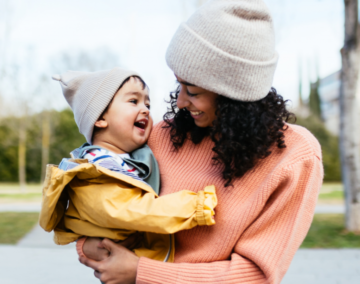 A mother wearing a winter hat holds her infant child and smiles.