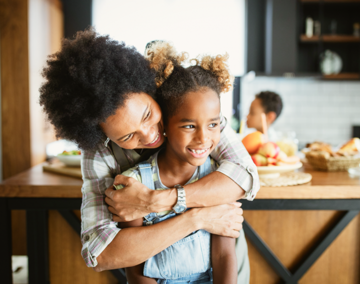 A mother hugs her daughter from behind as they stand in a kitchen.