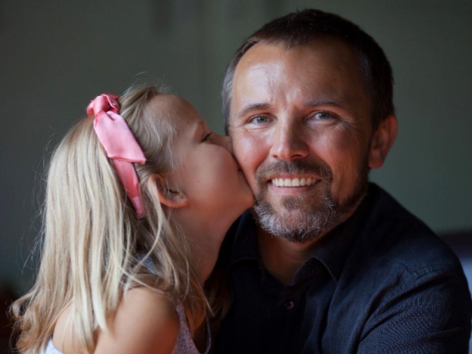 Daughter with a pink bow on her head kisses the cheek of her smiling father
