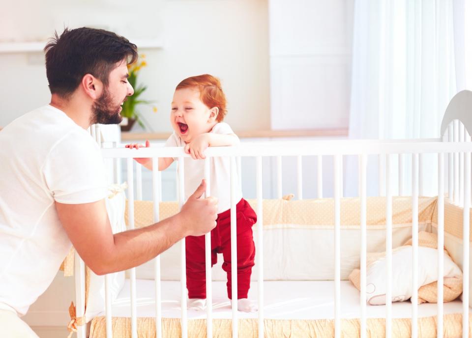 A father plays with his infant child on the side of the crib.