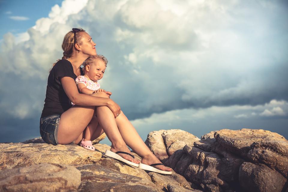Mom holds young daughter in her lap as they sit on top of a rock. 