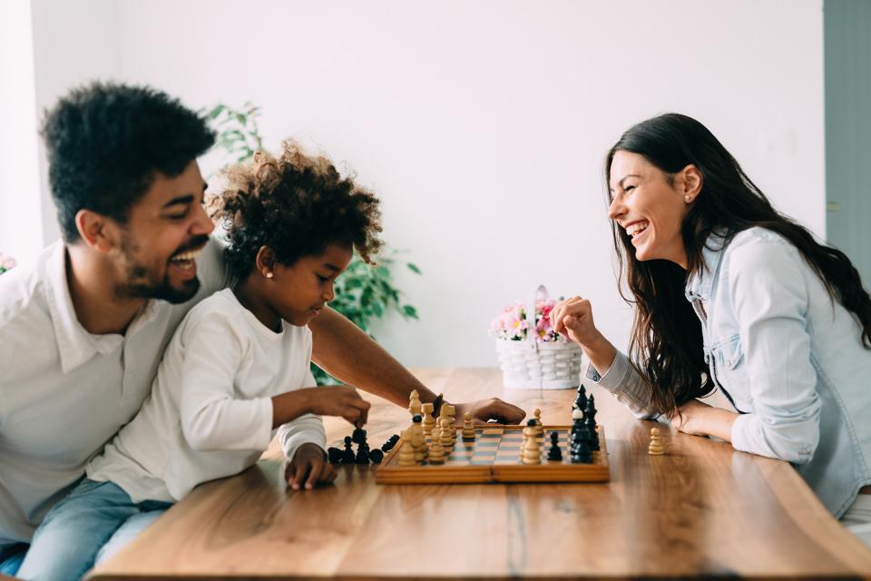 Co-parents play chess with their child together at a dining room table.