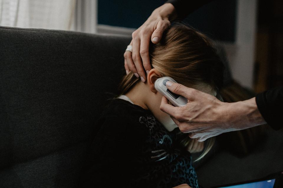 Parent takes temperature of child as they sit on the couch playing with a tablet