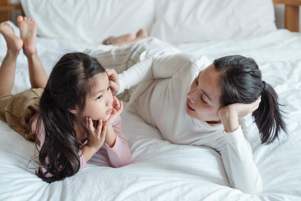 A mother and her daughter have a talk while lying next to each other on a bed with white sheets.