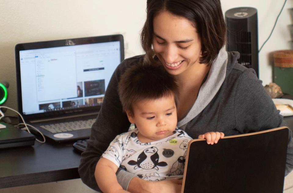 A mother sits at her desk with her child in her lap while she works on her laptop.