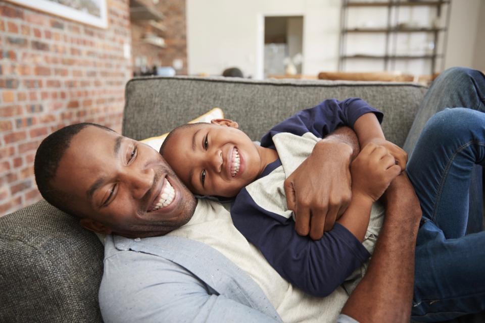 A child smiles as his father hugs him while laying on the couch.