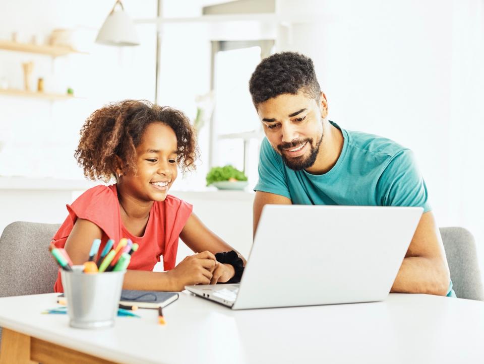 A father and daughter look at a laptop together and smile while sitting at a desk with a cup of pens nearby.