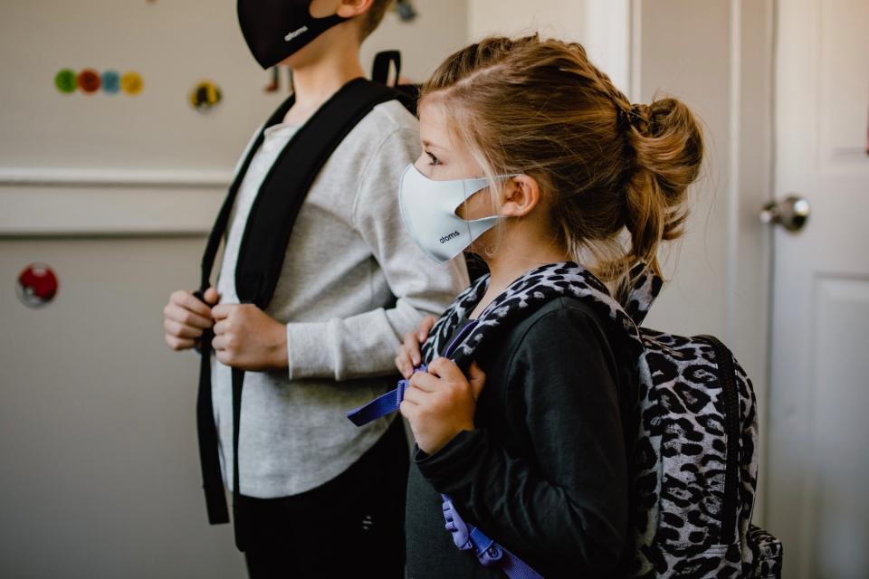 A young girl stands next to a boy while both wear face masks and backpacks to get ready for school.