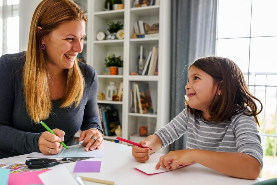 A woman and young girl sit together at a desk to write on cards and smile at each other