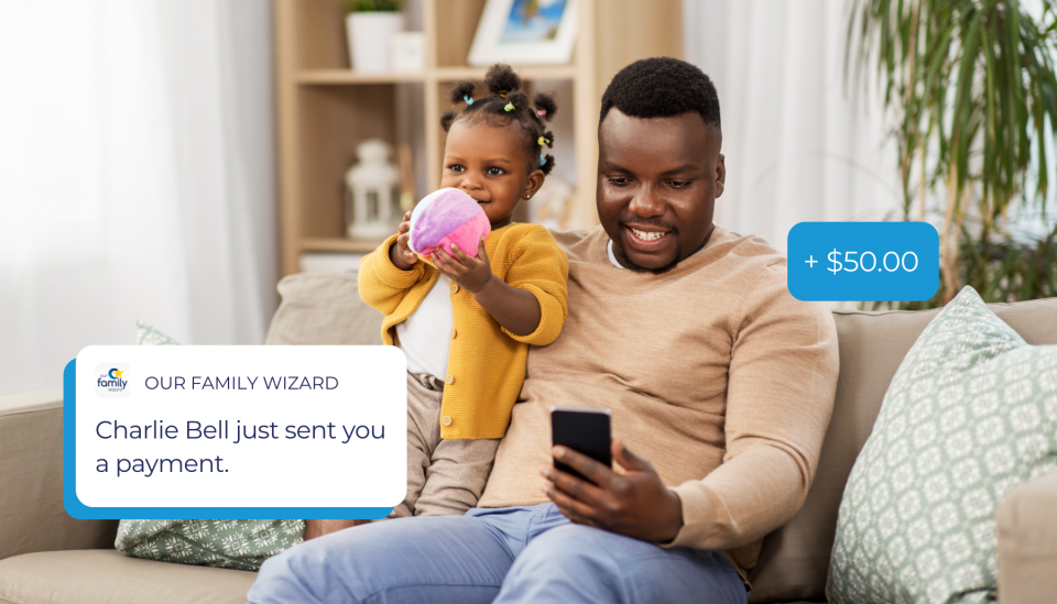 A young girl stands on the couch next to her father as he looks at his phone to check on an expense reimbursement.