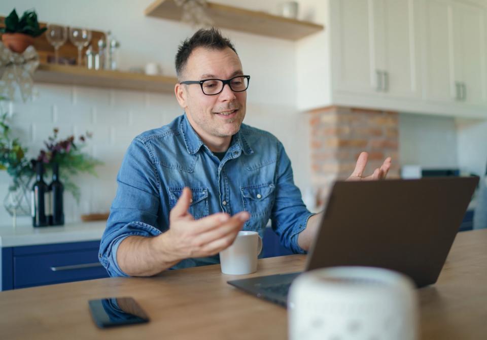 A man in a blue denim shirt and glasses sits at a kitchen table. He looks at the computer screen in front of him and holds his hands in the air.