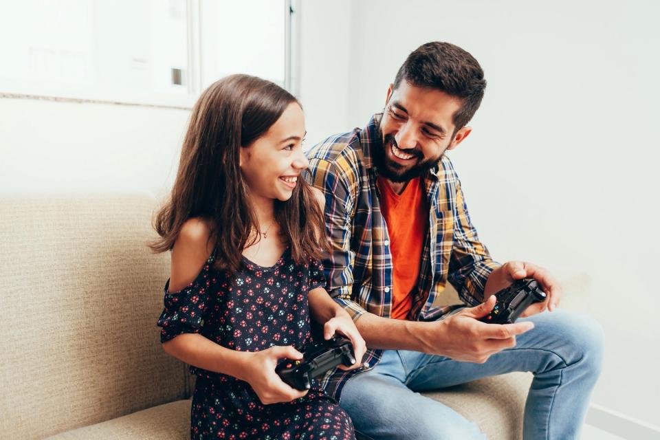A man with dark hair and a young girl with dark hair hold video game controllers. They are looking at each other and smiling.