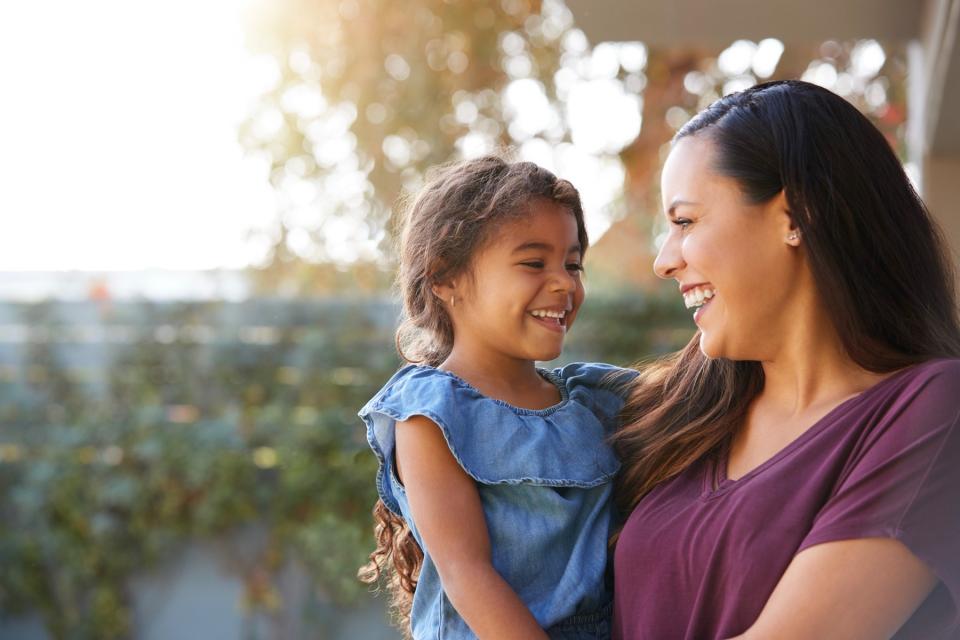 A woman holds a young girl while standing outside at sunset. They smile at each other.