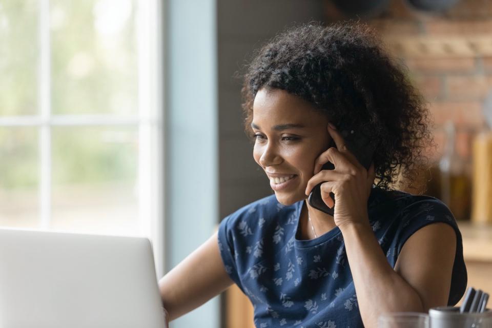 A woman in a blue shirt sits in front of a laptop, holds a cell phone to her ear, and smiles.