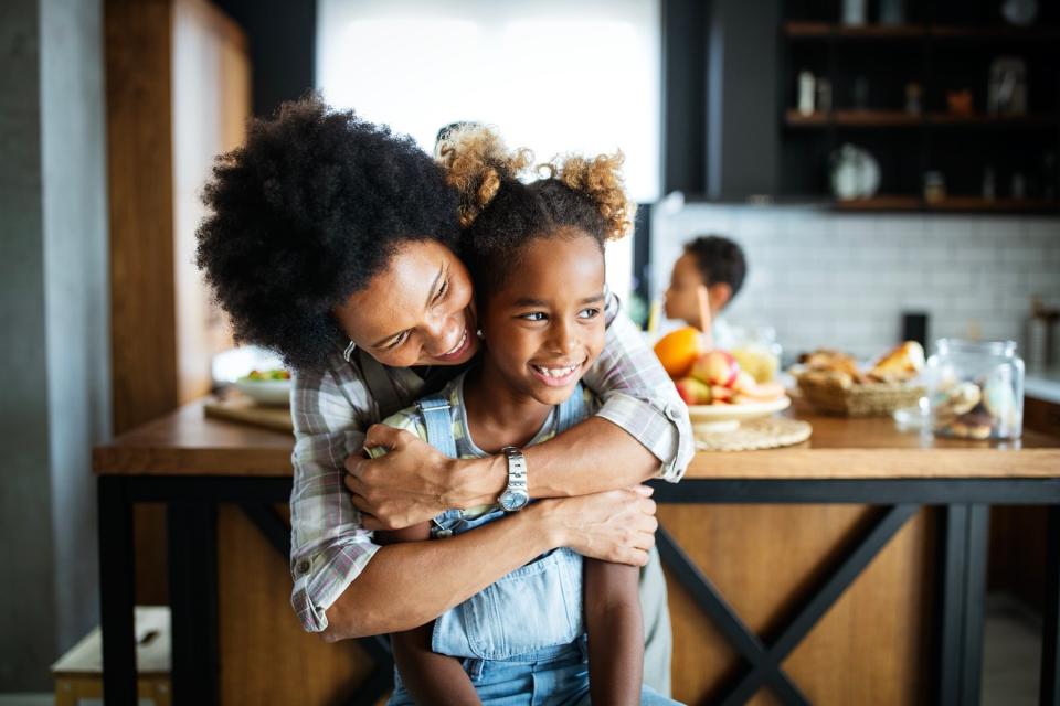 A mother and daughter stand in a kitchen and smile as the mother hugs the daughter from behind.