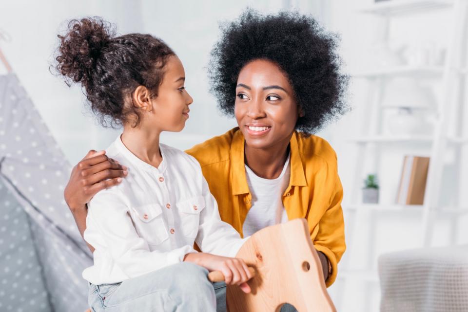 A young daughter turns her head and smiles at her mother, who is wearing a gold colored shirt.