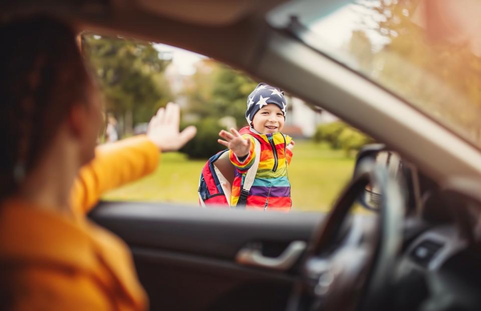 Young boy wearing a backpack waves to his mother outside her car on his way to school.