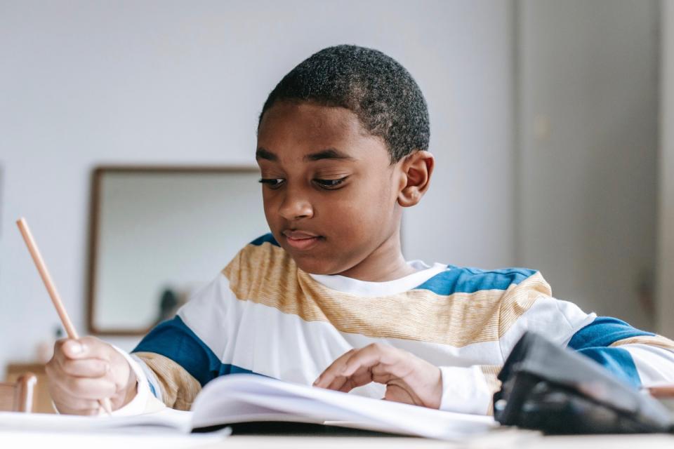 A boy in a striped shirt holds a pencil in one hand while writing in a notebook.