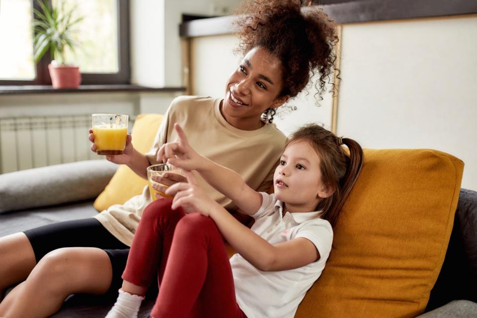 A female nanny sits on a couch and holds a glass of orange juice. A young girl sits next to her, pointing her finger forward.