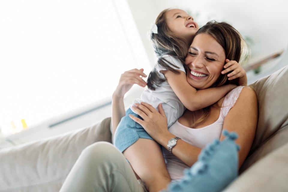 A young girl smiles and hugs her mother sitting on a couch.