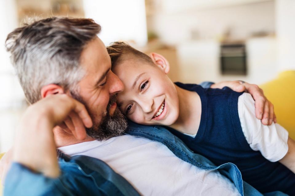 A boy and his father hug and smile while sitting on a couch.