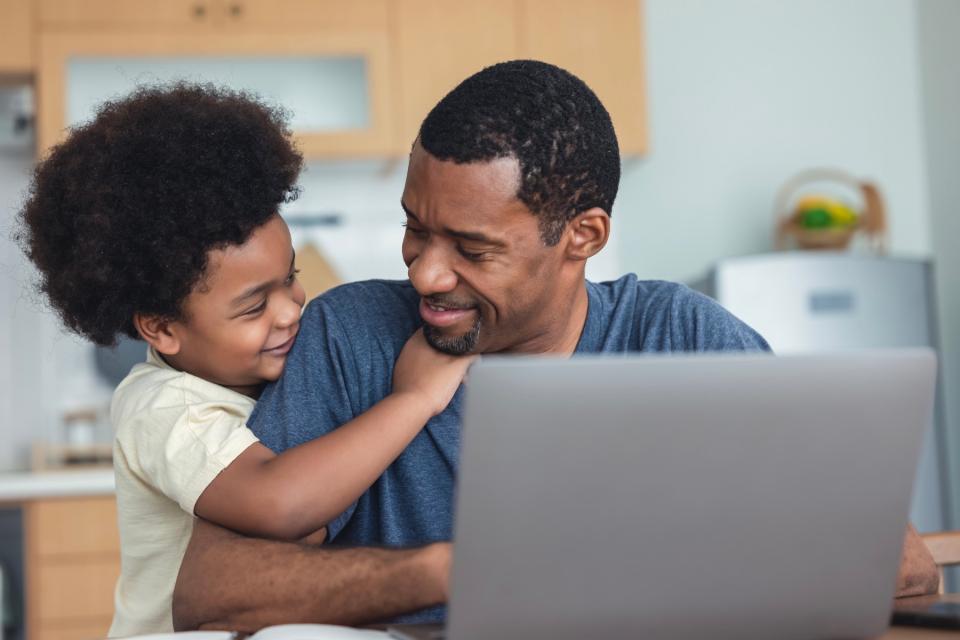 A young boy hugs his father from the size as Dad uses a computer.