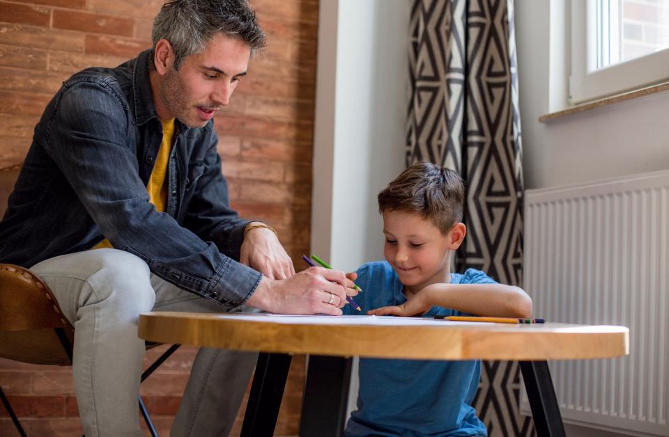 A father and son sit at a small table to draw pictures with colored pencils.