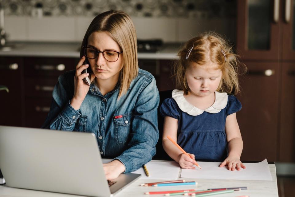 A mother works on a laptop while sitting at a kitchen table with her young daughter drawing on paper beside her.