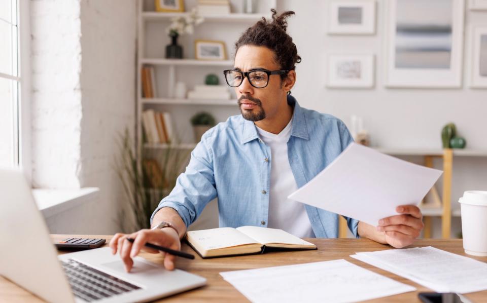 A man works on a laptop while also looking over papers in a living room.