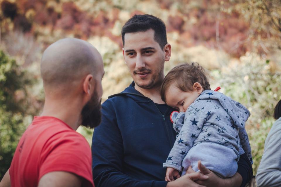 Two dads talking while one holds a sleeping baby.