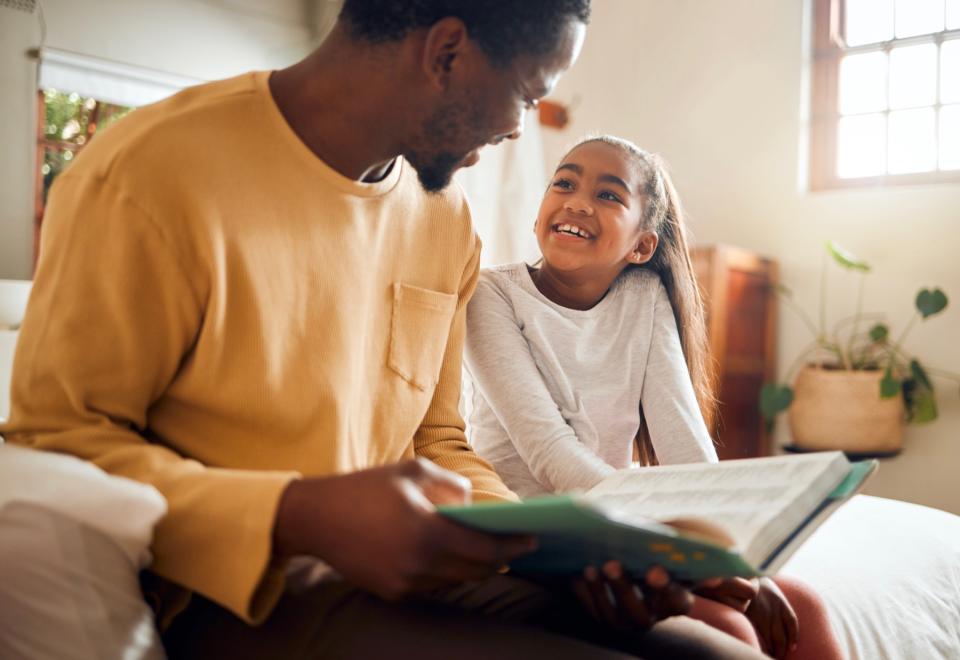 Father and daughter read together while sitting on a bed.