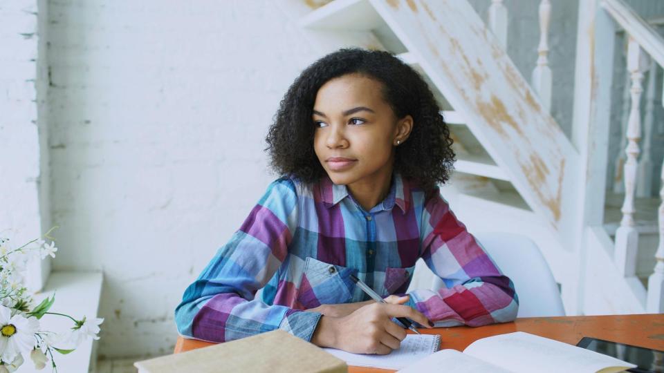 A teen girl sits at a table with her schoolwork, looking out the window
