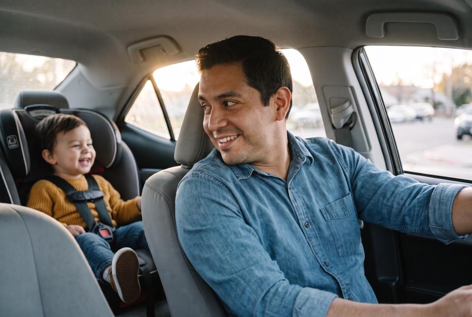 Smiling dad driving with his toddler buckled into a car seat in the back